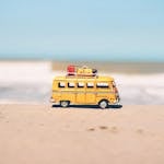 A vibrant yellow toy van sits on a sandy beach with an ocean backdrop, symbolizing fun travel adventures.