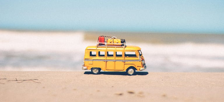 A vibrant yellow toy van sits on a sandy beach with an ocean backdrop, symbolizing fun travel adventures.