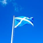 Scottish flag on a pole set against a vivid blue sky with scattered clouds.