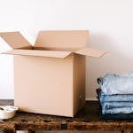 Open cardboard box on wooden table with folded jeans and packing tape nearby, ready for moving.