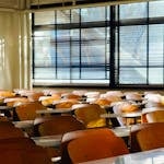 Sunlit empty classroom with wooden chairs and window blinds casting shadows across desks.