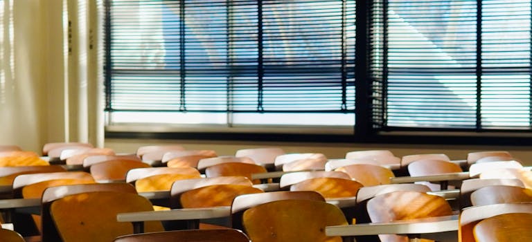 Sunlit empty classroom with wooden chairs and window blinds casting shadows across desks.