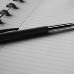 Black and white image of a pen resting on an open lined notebook, ideal for writing or note-taking themes.